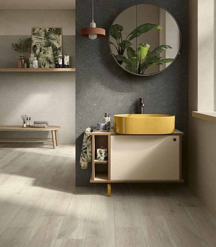 Contemporary bathroom featuring surfaces from the Surround Outline Soft, Draft and Borealis Inari collections, with a dark wall, yellow oval washbasin, floating vanity, large round mirror, decorative plants and light wood-effect flooring.