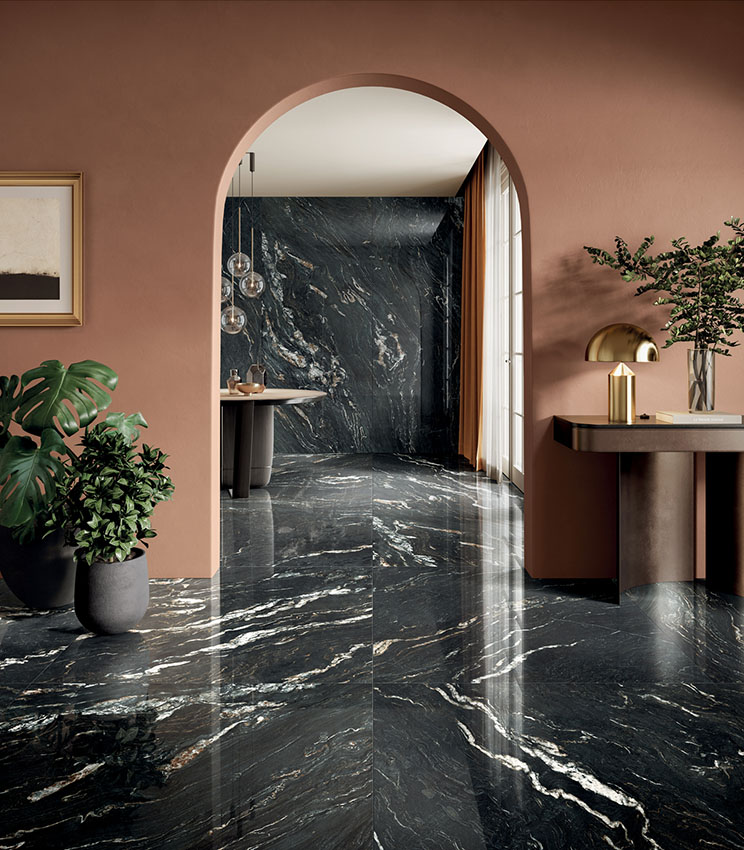Contemporary interior featuring surfaces from the Perpetual Belvedere Dark Lux collection, with large dark marble-effect slabs, light veining and a glossy finish. The space is completed by terracotta walls, minimalist furnishings, decorative plants, gold-toned lamps and an arched opening.