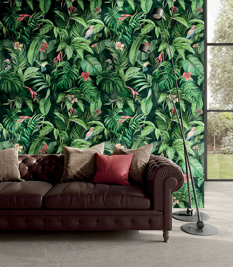 Contemporary living room featuring decorative wall covering from the Glam Vision collection, with a tropical pattern of leaves, flowers and birds, a dark leather sofa with neutral and burgundy cushions, black floor lamps and a large window overlooking the garden.