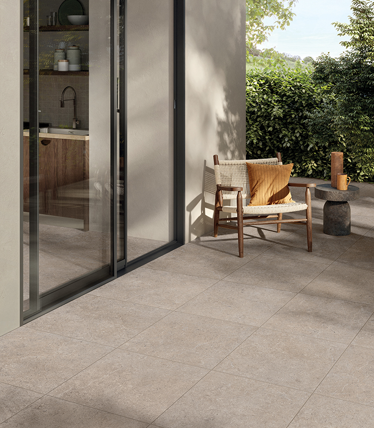 Contemporary terrace featuring flooring from the Surround Row collection, with large stone-effect slabs in beige tones, a wood and rope armchair, sculptural side table and lush greenery in the background. Large glass doors connect the outdoor space with the kitchen inside.