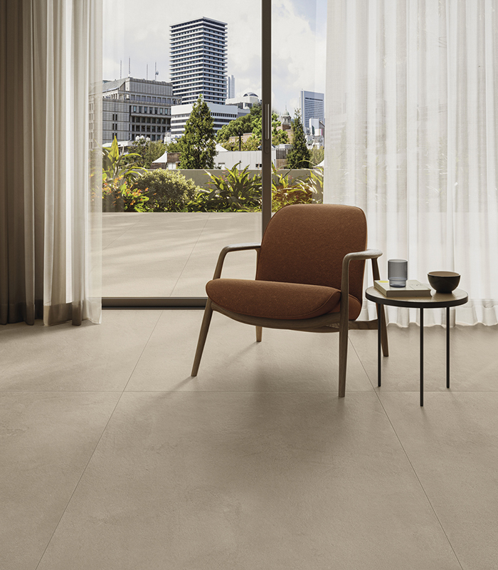 Partial view of a modern living room featuring an armchair and sliding door opening onto a terrace with a city view. Concrete-effect flooring by Panaria Ceramica, La Matière collection.