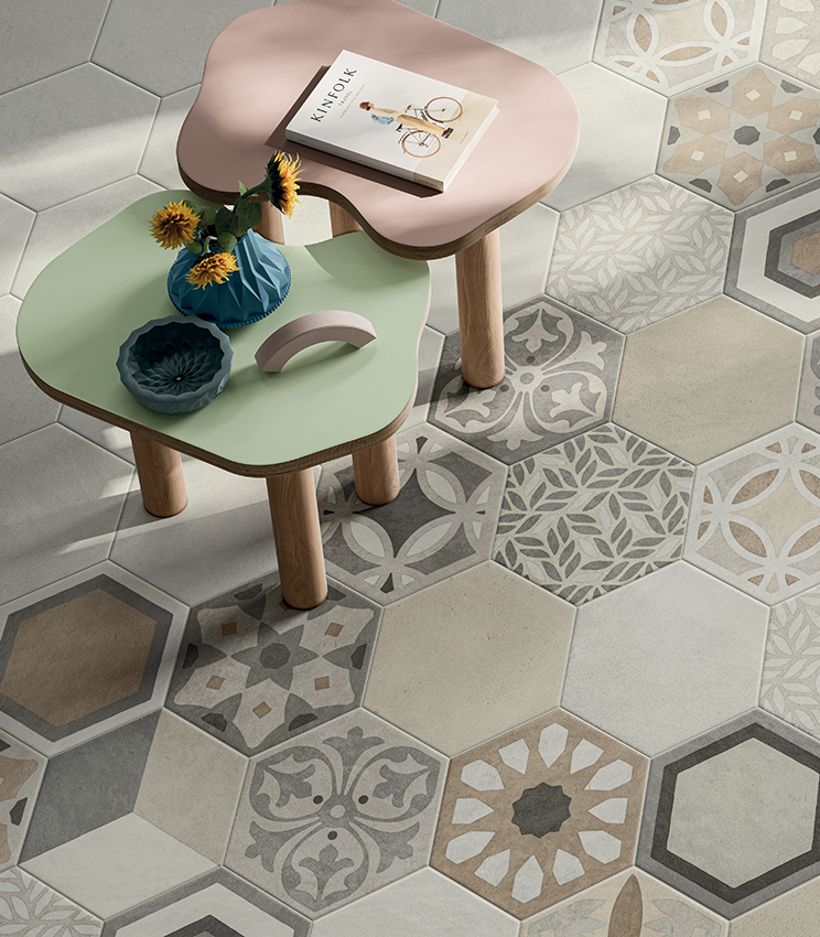 Detail of flooring featuring surfaces from the La Matière Hexa Talc Sable and Hexa Chic collections, with hexagonal tiles in neutral tones combined with geometric and floral decorative patterns. Two softly shaped green and pink tables complete the scene with vases and small decorative objects.