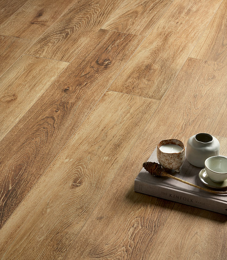Detail of flooring from the Kairos Ilios collection, featuring wood-effect surfaces with pronounced grain and warm, natural tones. The scene is completed by a book, a candle, a small vase and a ceramic cup.
