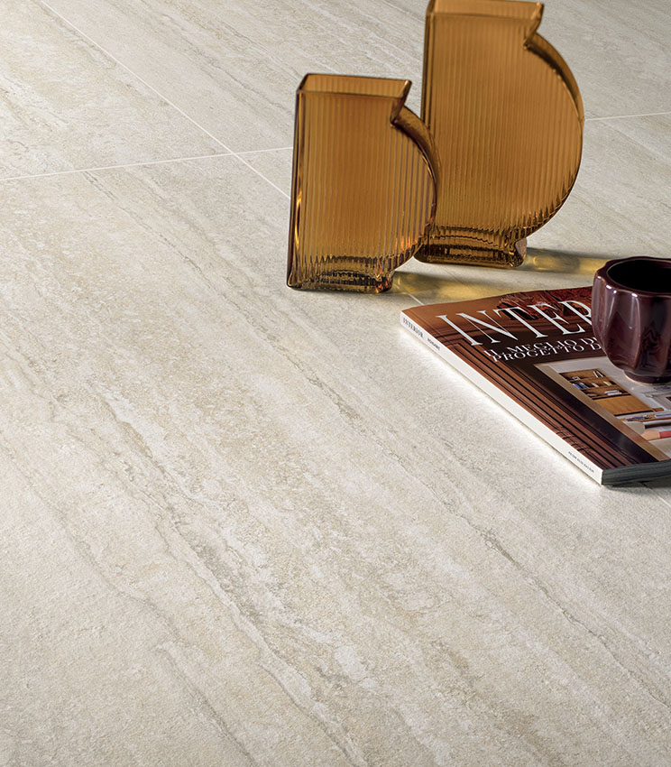 Detail of flooring from the Perpetual Travertino Ivory collection, featuring travertine-effect surfaces in light beige and ivory tones. The scene is completed by two amber glass vases, a magazine and a dark cup.