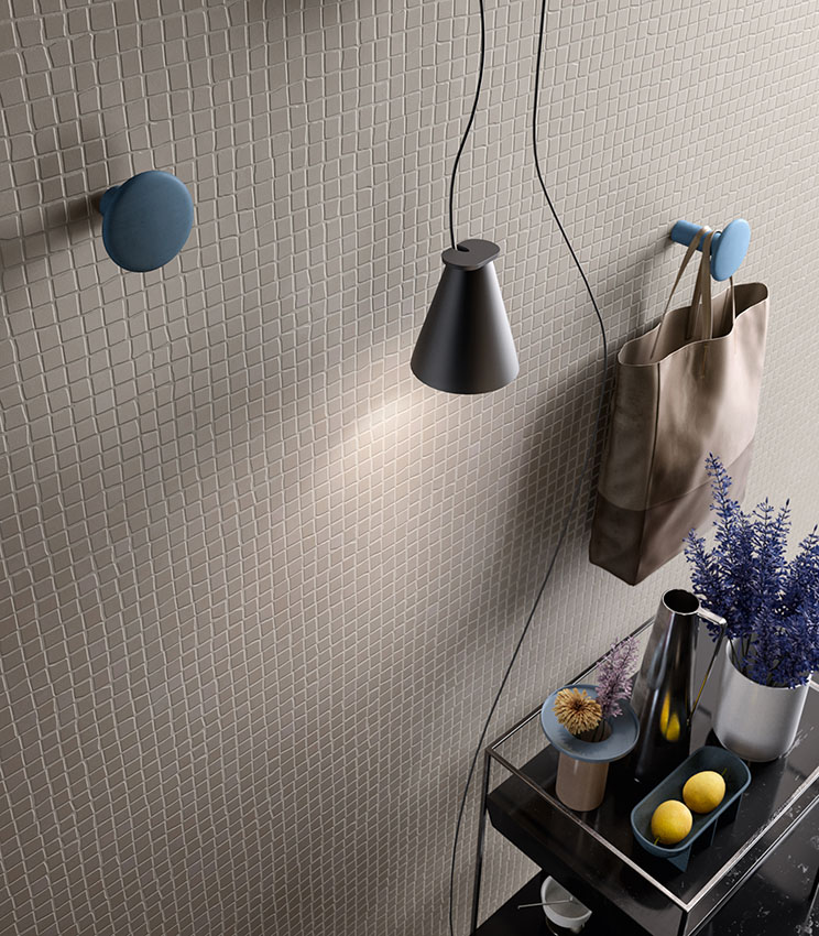 Detail of wall covering from the Even Dove Mosaico collection, featuring a regular pattern of small square tiles in light tones. The scene is completed by blue hooks, a black pendant lamp, a fabric bag and a console with vases, flowers and small decorative objects.
