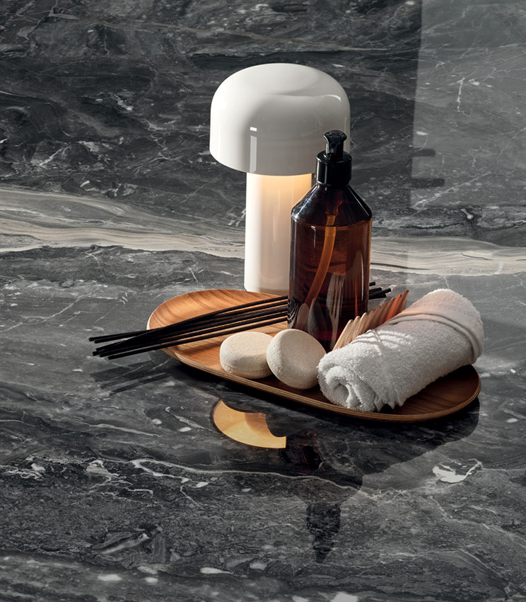 Detail of a surface from the Perpetual Orobico Blend Lux collection, featuring a dark marble effect with light veining and a glossy finish. The scene is completed by a wooden tray with a bottle, rolled towel, small accessories and a white table lamp.
