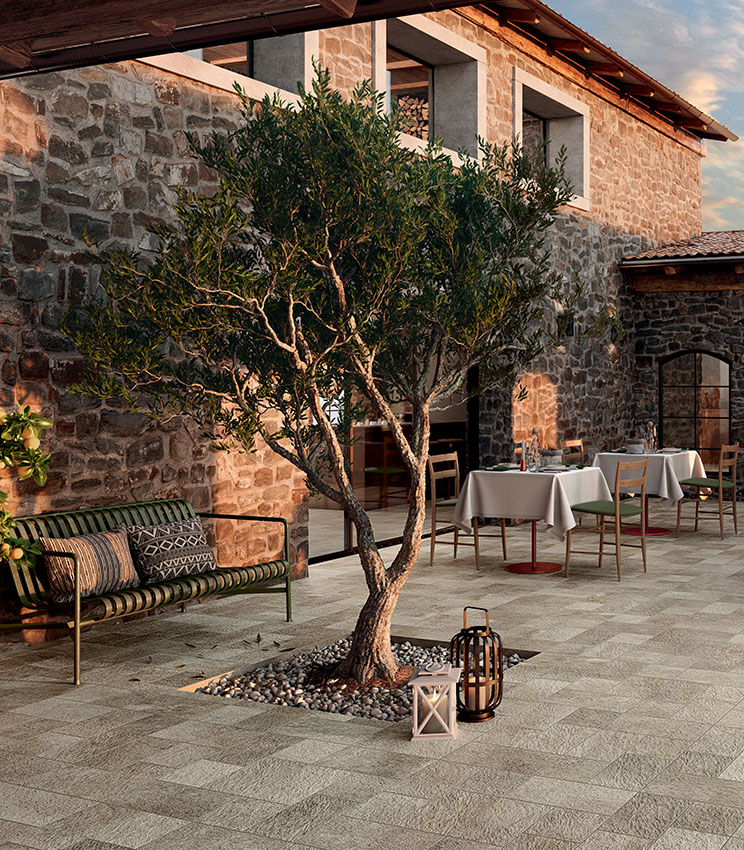 Restaurant terrace featuring flooring from the Pierre des Rêves Aurore collection, with stone-effect surfaces in natural tones. Surrounded by stone walls, the space is furnished with wooden tables and chairs, a bench, decorative lanterns and an olive tree at the centre.