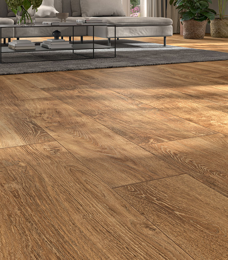 Contemporary living room featuring flooring from the Kairos Ilios collection, with warm wood-effect surfaces and pronounced grain. The space is furnished with a large grey sofa, a black metal coffee table, a dark rug and decorative plants lit by natural light.
