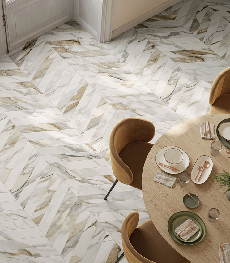 Contemporary dining room featuring flooring from the Perpetual Venato Gold Soft Chevron collection, with a white marble-effect chevron pattern and grey and golden veining. The space is furnished with a round light wood table, ochre upholstered chairs and a neutral-toned table setting.