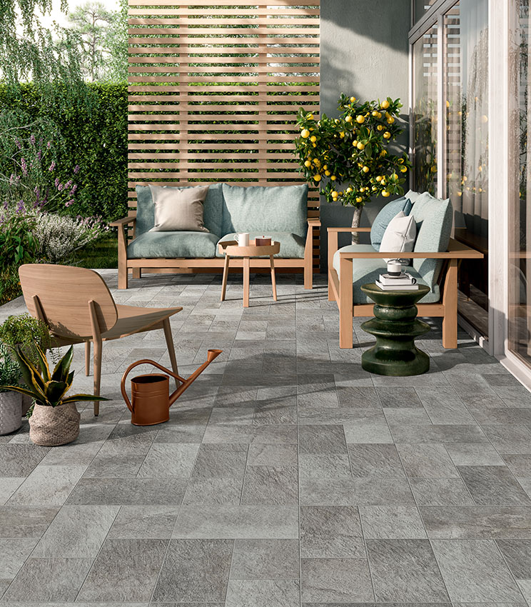 Contemporary terrace featuring flooring from the Pierre des Rêves Nuage collection, with grey stone-effect surfaces in modular formats. The space is furnished with wooden sofas and armchairs, sage green cushions, decorative side tables, potted plants and a citrus tree, surrounded by lush greenery.