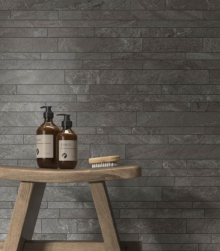 Scorcio frontale di parete rivestita con listelli multiformato di gres porcellanato effetto pietra posati a correre. Contro la parete si trova uno sgabello di legno sul quale poggiano prodotti di igiene personale