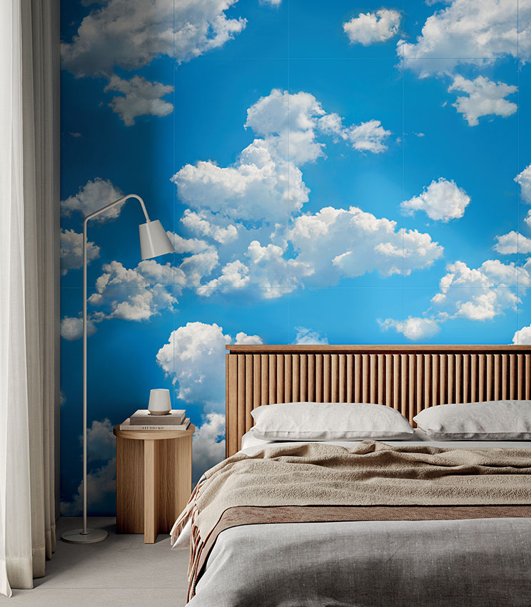 Contemporary bedroom featuring decorative wall covering from the Glam Flow collection, depicting a blue sky with large white clouds. The room is furnished with a bed with a fluted wooden headboard, neutral-toned bedding, a wooden bedside table and a white floor lamp.