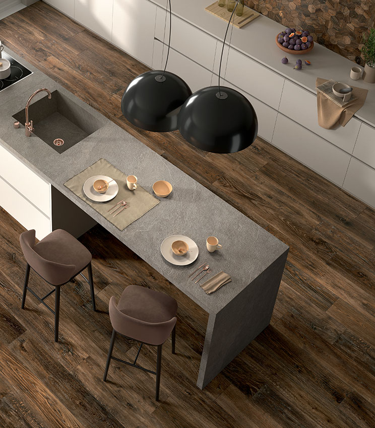 Top view of a modern kitchen with a dinette island covered with light grey stone-effect stoneware slabs and dark wood-effect porcelain stoneware floor tiles.