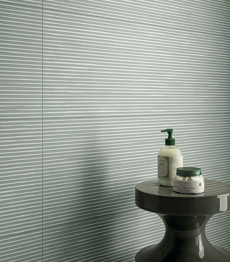 Detail of a contemporary bathroom featuring wall covering from the Lullaby Linear Green collection, with a light green surface and horizontal linear pattern. The scene is completed by a dark sculptural side table with bottles and small bathroom products.