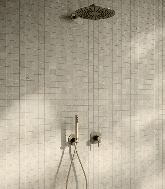 Detail of a shower wall clad with beige stone-effect ceramic mosaic, featuring minimalist light gold fittings and a matte finish.