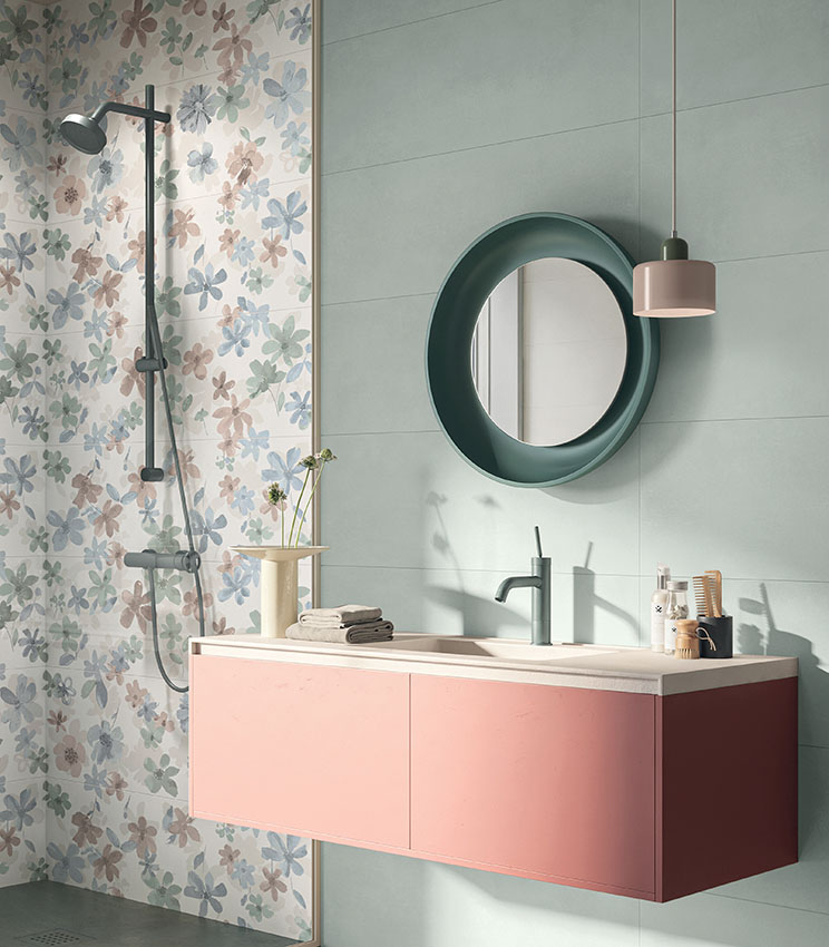 Contemporary bathroom featuring surfaces from the Lullaby Silent Green, Florè and Borealis Abisko collections, with light green wall coverings and a floral feature wall in pastel tones. The space includes a pink floating vanity, a dark green round mirror, coordinated tapware, a pendant light and a shower area with a black shower column.