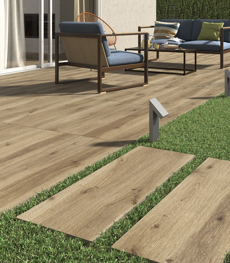 View of a patio with a modern outdoor seating area comprising a metal armchair and sofa. Floor in 20 mm wood-effect porcelain stoneware laid with adhesive. In the foreground, an outdoor ceramic plank pathway begins, dry-laid on grass | Borealis collection by Panaria Ceramica