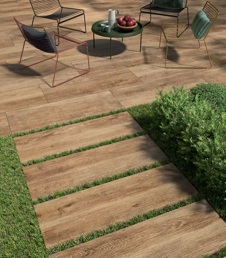 View of a patio with modern wire-frame chairs and a 20 mm wood-effect porcelain stoneware floor laid with adhesive. In the foreground, an outdoor ceramic plank pathway begins, dry-laid on grass | Kairos collection by Panaria Ceramica