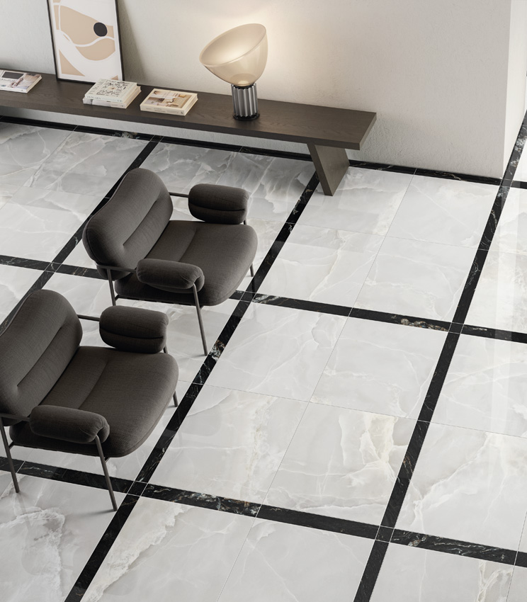 Elevated view of a large, bright living area with a black and white marble-effect porcelain stoneware floor laid in a framed carpet pattern, featuring a pair of dark armchairs and a bench unit leaning against a white partition wall.