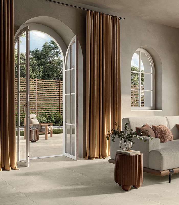 Scorcio di elegante living neoclassicheggiante, con porta-finestra ad arco a tutto sesto aperta su patio, pavimento in gres porcellanato indoor-outdoor effetto pietra color sabbia.