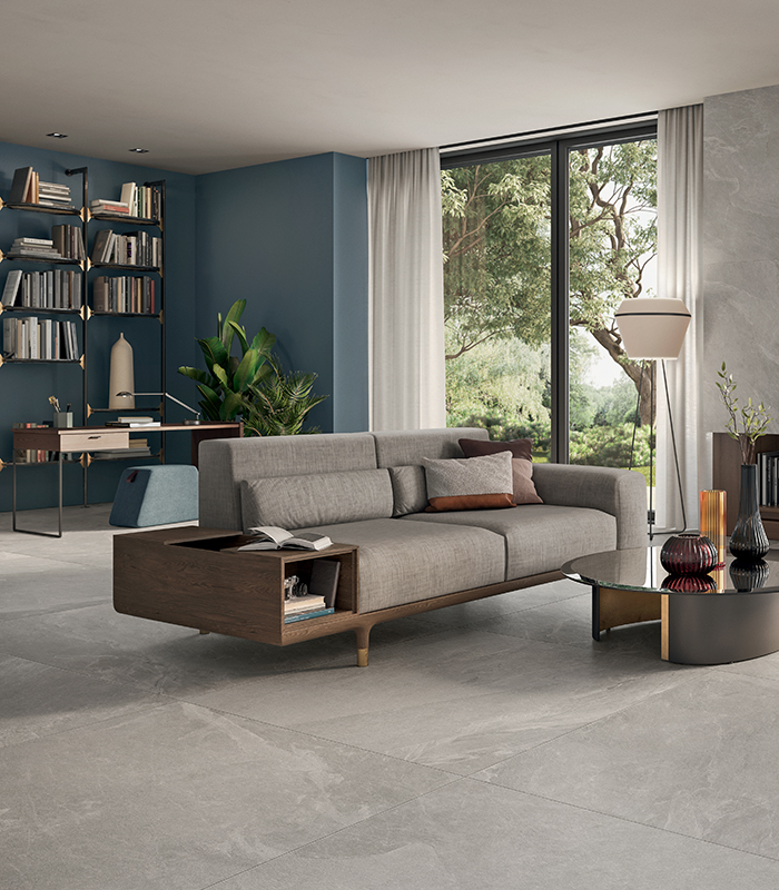 Living contemporaneo e minimalista dalle tonalità neutre con accenti di blu per la parete dell’angolo di fondo dedicato a un piccolo studiolo e di arancione per una poltrona. Pavimento e rivestimento in grandi lastre di gres porcellanato sottile effetto pietra grigia.