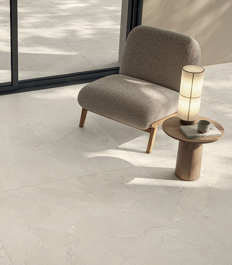 Bright modern living area with an upholstered armchair, wooden side table and light stone-effect porcelain stoneware flooring, extending seamlessly outdoors.