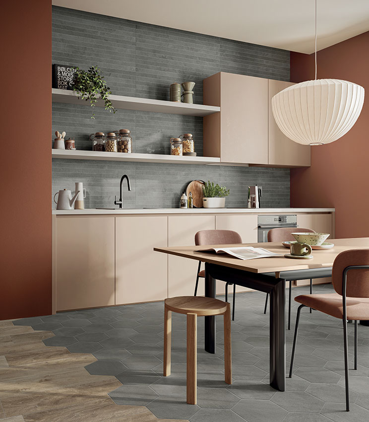 Contemporary kitchen with grey linear wall tiles and geometric flooring, furnished with beige cabinetry, terracotta walls, a dining table with upholstered chairs and a large white pendant lamp.