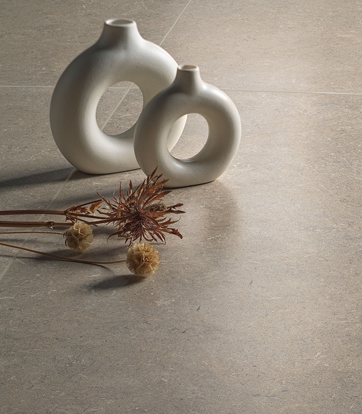 Detail of flooring from the Surround Row Soft collection, in a beige stone-effect finish, styled with two light sculptural vases and dried flower arrangements.
