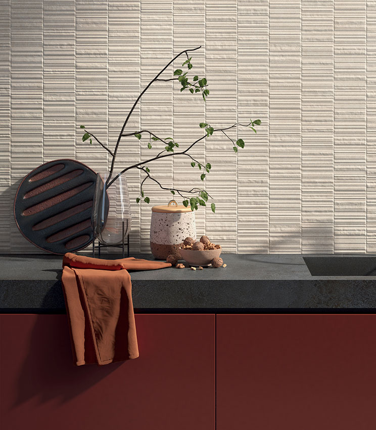 Contemporary kitchen featuring Iqoniqa Brix Skin wall tiles, with a light textured wall, dark worktop, terracotta cabinetry and a decorative arrangement of vases, green branches and ceramic objects.