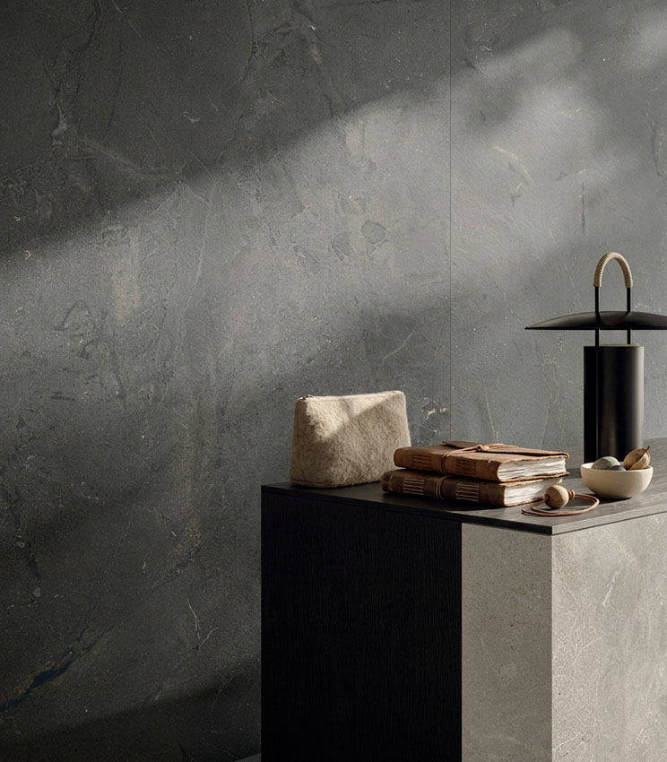 Detail of surfaces from the Revel Night and Revel Ground collections, featuring large stone-effect slabs in dark grey and beige tones. The scene is completed by a minimalist console, books, small decorative objects and a black lamp, highlighted by soft natural light.