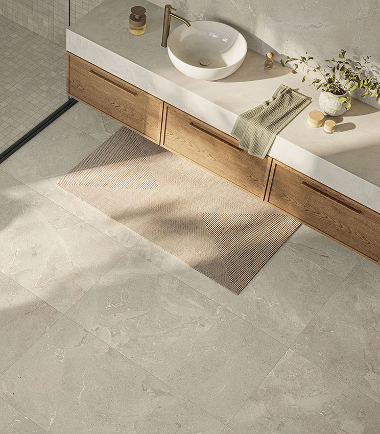 Contemporary bathroom featuring surfaces from the Revel Dune, Mosaico Dune and Workshop Jade collections, with light stone-effect flooring and wall coverings. The space includes a long wooden vanity, a light countertop with a white vessel sink, gold-toned tapware, a geometric rug and a shower with coordinated mosaic tiles.