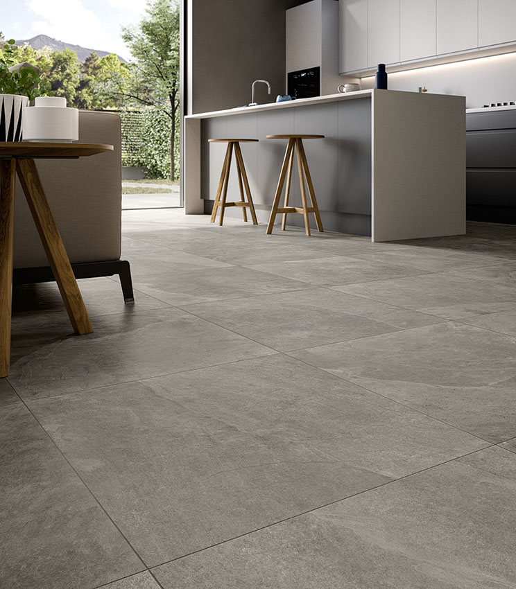 Open-plan living and kitchen with white island, grey stone-effect porcelain floor tiles from the Frame Peak collection, sofa and wooden side table, sliding glass door opening onto a garden with mountain view.