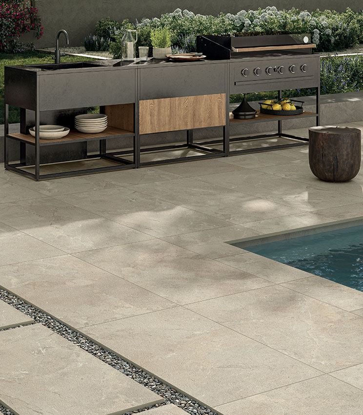 Poolside area with stone-effect porcelain stoneware flooring, outdoor kitchen and contemporary furnishings, creating an elegant, durable and functional space.